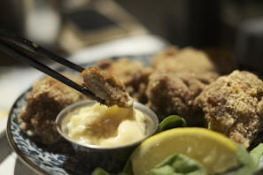 Chopsticks holding a piece of breaded, fried chicken above a small dish of creamy dipping sauce. Several other pieces of fried chicken are arranged on a decorative plate. A wedge of lemon and leafy greens are visible in the foreground, adding freshness to the presentation.