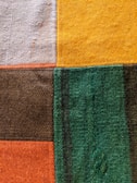 A close-up view of a textured fabric featuring a patchwork design composed of five rectangular sections in different colors: beige, yellow, dark brown, green, and orange. The fabric surface displays a woven texture with visible fibers and faint imperfections.