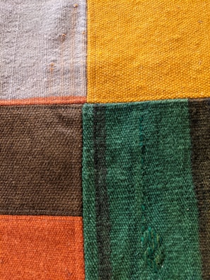 A close-up view of a textured fabric featuring a patchwork design composed of five rectangular sections in different colors: beige, yellow, dark brown, green, and orange. The fabric surface displays a woven texture with visible fibers and faint imperfections.