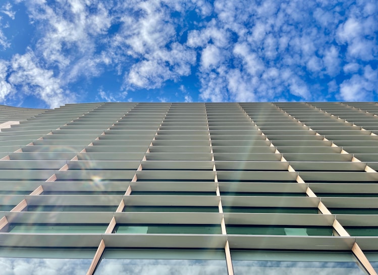A modern architectural facade with horizontal lines and large glass windows, set against a backdrop of a bright blue sky filled with scattered clouds.
