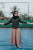 A person stands in front of a tennis court net wearing a dark green sweater, a black headscarf, and a long pink skirt. In the background, there are palm trees and part of a building visibly behind the fence.