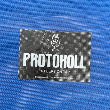 A black rectangular card with white text and a simple line drawing of a beer glass. It reads 'PROTOKOLL' in bold letters, followed by '24 BEERS ON TAP' in smaller font. The card is placed on a textured blue background.