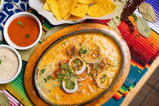 An oval cast iron dish filled with a cheesy, rich dip garnished with slices of red onion and green herbs. The dish is surrounded by an assortment of culinary elements, including a bowl of red chili powder, a bowl of sesame seeds, yellow corn tortilla chips, and bay leaves. The setting features a vibrant, colorful woven tablecloth with stripes in red, yellow, blue, and green.