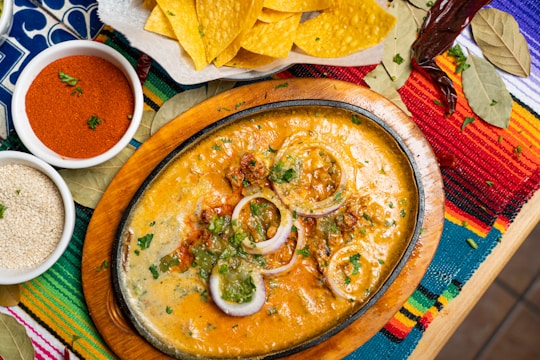 An oval cast iron dish filled with a cheesy, rich dip garnished with slices of red onion and green herbs. The dish is surrounded by an assortment of culinary elements, including a bowl of red chili powder, a bowl of sesame seeds, yellow corn tortilla chips, and bay leaves. The setting features a vibrant, colorful woven tablecloth with stripes in red, yellow, blue, and green.