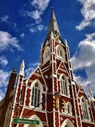 A red brick church with white accents and a tall, pointed spire set against a blue sky with scattered clouds. The building features Gothic-style architecture with arched windows and a clock on the steeple. In the foreground, a street sign indicates 'Manhattan Av' and a traffic light is visible.