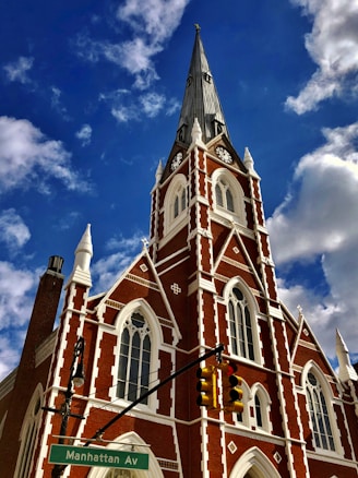A red brick church with white accents and a tall, pointed spire set against a blue sky with scattered clouds. The building features Gothic-style architecture with arched windows and a clock on the steeple. In the foreground, a street sign indicates 'Manhattan Av' and a traffic light is visible.