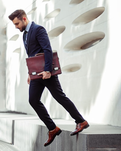 A professionally dressed man in a suit is stepping down from a ledge while holding a briefcase. The background features a modern architectural wall with circular cutouts.
