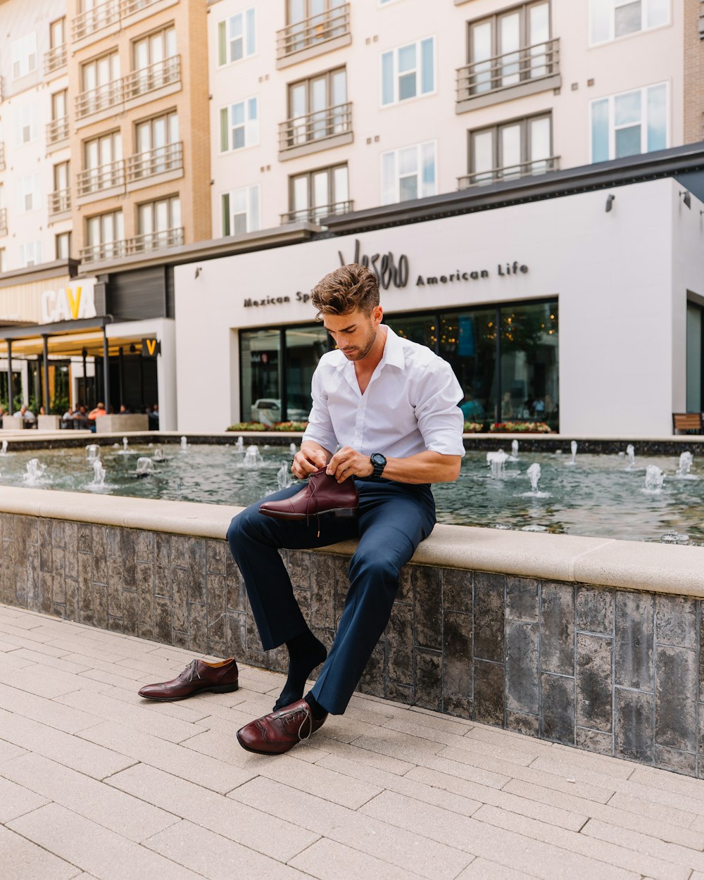 man in white dress shirt and blue denim jeans sitting on concrete bench near water fountain