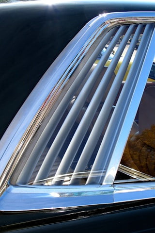A close-up view of a car window with several vertical metal slats inside the window frame. The chrome trim of the window reflects light, enhancing its shine. The interior of the car is partially visible through the slats, and there are subtle reflections on the glass surface.