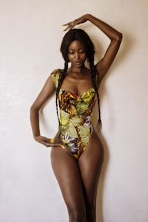A person stands against a plain background, wearing a colorful, tropical-patterned swimsuit. Their long hair is styled in braids, and their pose is elegant with one arm raised and the other hand resting near their waist.