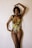 A person stands against a plain background, wearing a colorful, tropical-patterned swimsuit. Their long hair is styled in braids, and their pose is elegant with one arm raised and the other hand resting near their waist.