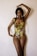 A person stands against a plain background, wearing a colorful, tropical-patterned swimsuit. Their long hair is styled in braids, and their pose is elegant with one arm raised and the other hand resting near their waist.