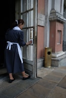 A woman in a dark uniform with a white apron and hair ribbon is cleaning the handle of a door. The setting appears to be the entrance to an old building with stone walls and a metal grate on the ground. A brass-finished metal container is positioned near the entrance.