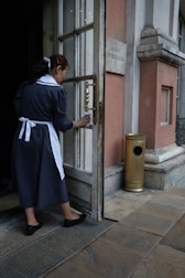 A woman in a dark uniform with a white apron and hair ribbon is cleaning the handle of a door. The setting appears to be the entrance to an old building with stone walls and a metal grate on the ground. A brass-finished metal container is positioned near the entrance.