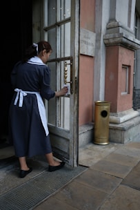 A woman in a dark uniform with a white apron and hair ribbon is cleaning the handle of a door. The setting appears to be the entrance to an old building with stone walls and a metal grate on the ground. A brass-finished metal container is positioned near the entrance.