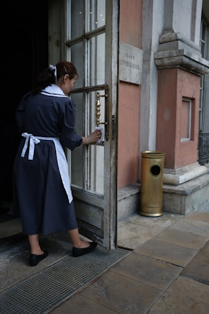 A woman in a dark uniform with a white apron and hair ribbon is cleaning the handle of a door. The setting appears to be the entrance to an old building with stone walls and a metal grate on the ground. A brass-finished metal container is positioned near the entrance.