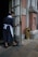 A woman in a dark uniform with a white apron and hair ribbon is cleaning the handle of a door. The setting appears to be the entrance to an old building with stone walls and a metal grate on the ground. A brass-finished metal container is positioned near the entrance.