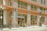 A modern urban coffee shop with a large glass facade and tall street lamps. The entrance is marked by a vertical sign that reads 'Coffee Roaster & Wine Bar'. A traffic light and a street sign for Washington Street are visible. People and pets stroll along the sidewalk, adding life to the scene.