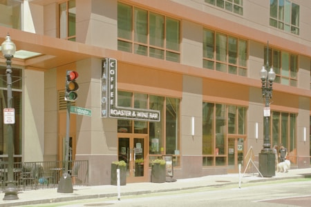 A modern urban coffee shop with a large glass facade and tall street lamps. The entrance is marked by a vertical sign that reads 'Coffee Roaster & Wine Bar'. A traffic light and a street sign for Washington Street are visible. People and pets stroll along the sidewalk, adding life to the scene.