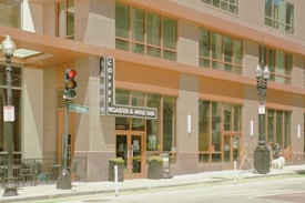 A modern urban coffee shop with a large glass facade and tall street lamps. The entrance is marked by a vertical sign that reads 'Coffee Roaster & Wine Bar'. A traffic light and a street sign for Washington Street are visible. People and pets stroll along the sidewalk, adding life to the scene.