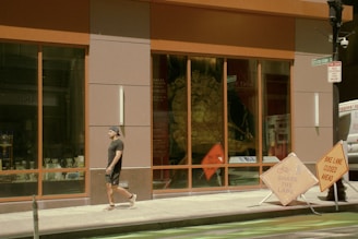 A man dressed casually in dark clothing walks along a city sidewalk past a large store window displaying various items. Bright orange construction signs are placed nearby, indicating a closure of a bike lane. The window reflects a mix of outdoor and indoor elements, creating a dynamic urban atmosphere.