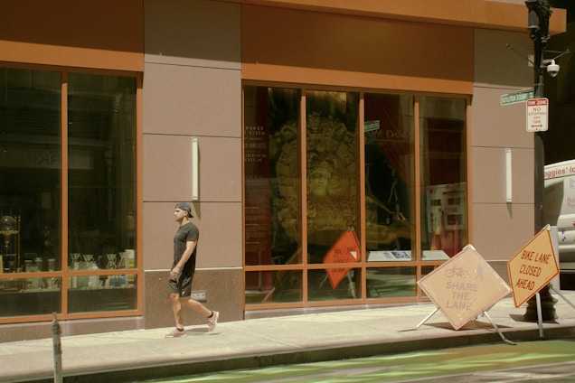 A man dressed casually in dark clothing walks along a city sidewalk past a large store window displaying various items. Bright orange construction signs are placed nearby, indicating a closure of a bike lane. The window reflects a mix of outdoor and indoor elements, creating a dynamic urban atmosphere.