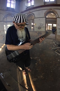 An elderly man with a long white beard is playing a bass guitar in a dilapidated building with peeling paint and graffiti on the walls. He is wearing a black shirt, pants, sandals, and a striped bandana, along with round sunglasses. Sunlight enters through broken windows, casting patterns on the dusty floor.