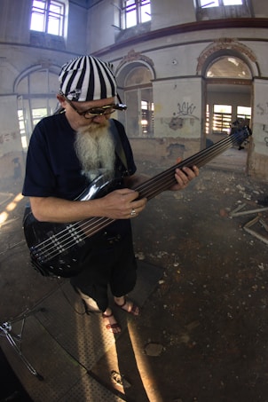 An elderly man with a long white beard is playing a bass guitar in a dilapidated building with peeling paint and graffiti on the walls. He is wearing a black shirt, pants, sandals, and a striped bandana, along with round sunglasses. Sunlight enters through broken windows, casting patterns on the dusty floor.