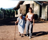 An elderly woman using a cane is supported by a younger woman as they walk together outside. They are on a dirt path near a rustic building with wooden doors. In the background, there are trees and a view of hills or mountains.