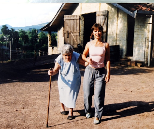 An elderly woman using a cane is supported by a younger woman as they walk together outside. They are on a dirt path near a rustic building with wooden doors. In the background, there are trees and a view of hills or mountains.