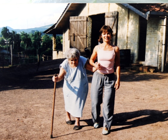 An elderly woman using a cane is supported by a younger woman as they walk together outside. They are on a dirt path near a rustic building with wooden doors. In the background, there are trees and a view of hills or mountains.