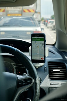 The interior of a car is depicted, focusing on the dashboard area. A smartphone is mounted on the dashboard, displaying a navigation app with a route, instructing to stay on Route 33. The steering wheel is visible on the left, and there are buttons for cruise control. Outside the car, traffic can be seen with several vehicles on the road.