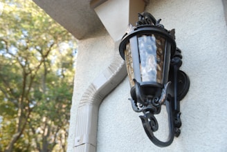 An ornate black metal lantern is mounted on a textured wall. The glass panes of the lantern have a bubbled texture, reflecting light. A beige drainpipe runs alongside the lantern, curving with the wall’s architecture. In the background, blurred out-of-focus trees with green foliage are visible.