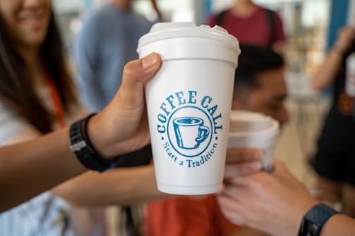 A group of people holding white coffee cups with a blue logo that reads 'Coffee Call Start a Tradition.' Some individuals are slightly blurred in the background, suggesting a social and casual setting.