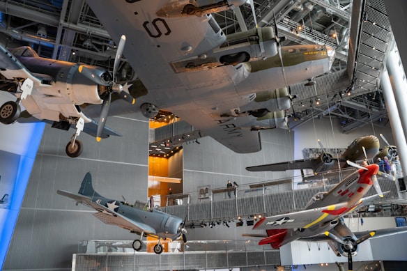 Several vintage military aircraft are suspended from the ceiling of a large, modern museum. The planes vary in size and style, featuring propellers and painted in different colors and markings. The setting is well-lit with visible metal beams and background lighting, providing a view of the museum's interior structure.