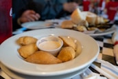 A plate of small, golden-brown empanadas arranged around a small cup of dipping sauce sits on a checkered tablecloth. In the background, there are blurred images of a person, red cups, and a plate with sliced bread and other food items.