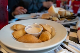 A plate of small, golden-brown empanadas arranged around a small cup of dipping sauce sits on a checkered tablecloth. In the background, there are blurred images of a person, red cups, and a plate with sliced bread and other food items.