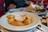 A plate of small, golden-brown empanadas arranged around a small cup of dipping sauce sits on a checkered tablecloth. In the background, there are blurred images of a person, red cups, and a plate with sliced bread and other food items.