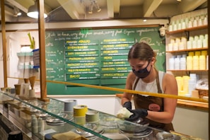 A woman wearing a face mask is inside an ice cream shop, scooping ice cream behind a glass display. Behind her is a chalkboard menu displaying various ice cream flavors. The interior has shelves filled with stacked cups in various pastel colors.