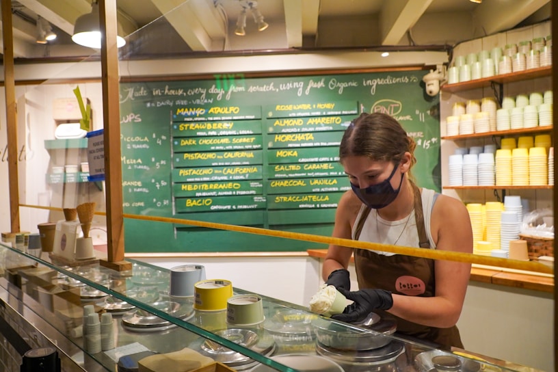 A woman wearing a face mask is inside an ice cream shop, scooping ice cream behind a glass display. Behind her is a chalkboard menu displaying various ice cream flavors. The interior has shelves filled with stacked cups in various pastel colors.