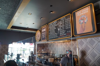 A modern ice cream shop interior featuring a large menu board listing various flavors and prices. The decor includes geometric patterned walls, stylish lighting, and a playful sign with an illustration of flowers in an ice cream cone accompanied by the text 'EAT YOUR FLOWERS'. A masked staff member is visible behind a glass counter displaying containers, likely containing ice cream or toppings.