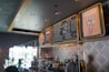 A modern ice cream shop interior featuring a large menu board listing various flavors and prices. The decor includes geometric patterned walls, stylish lighting, and a playful sign with an illustration of flowers in an ice cream cone accompanied by the text 'EAT YOUR FLOWERS'. A masked staff member is visible behind a glass counter displaying containers, likely containing ice cream or toppings.