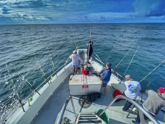 Several people are on a fishing boat in the open sea. The boat is equipped with multiple fishing rods, and the expansive ocean surrounds them under a cloudy sky. The group appears to be engaged in fishing activities with various gear visible on the deck.