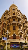 A historic architectural building featuring ornate balconies with decorative railings and arched windows. The structure is multi-storied with a distinct dome on top. Beneath the building is a pharmacy with a colorful sign in both Arabic and English.