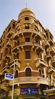 A historic architectural building featuring ornate balconies with decorative railings and arched windows. The structure is multi-storied with a distinct dome on top. Beneath the building is a pharmacy with a colorful sign in both Arabic and English.