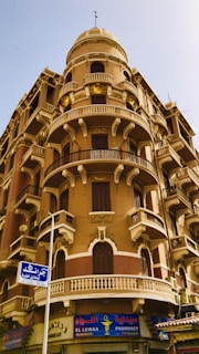 A historic architectural building featuring ornate balconies with decorative railings and arched windows. The structure is multi-storied with a distinct dome on top. Beneath the building is a pharmacy with a colorful sign in both Arabic and English.