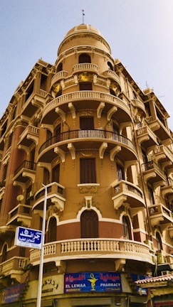 A historic architectural building featuring ornate balconies with decorative railings and arched windows. The structure is multi-storied with a distinct dome on top. Beneath the building is a pharmacy with a colorful sign in both Arabic and English.