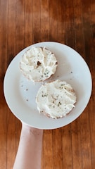 Two halves of a bagel are covered with cream cheese and sprinkled with herbs, placed on a white plate. The plate is being held up by a hand over a wooden floor, creating a rustic and homemade atmosphere.