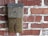 A vintage metal mailbox labeled 'MAIL' is mounted on a red brick wall. The mailbox appears weathered and aged, with a rustic patina. The brick wall has an uneven surface with visible mortar between the bricks.