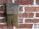 A vintage metal mailbox labeled 'MAIL' is mounted on a red brick wall. The mailbox appears weathered and aged, with a rustic patina. The brick wall has an uneven surface with visible mortar between the bricks.